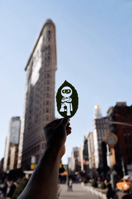 A hand holds up a leaf cutout featuring a white robot character silhouette, silhouetted against the iconic Flatiron Building in a busy city street.
