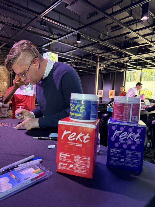 The image features a man sitting at a table with two boxes of Rekt champagne, one red and one blue. The man appears to be examining or testing the contents of the boxes. There are also some pens and markers on the table next to the boxes. In the background, there is another person who might be involved in the process or simply observing the scene.