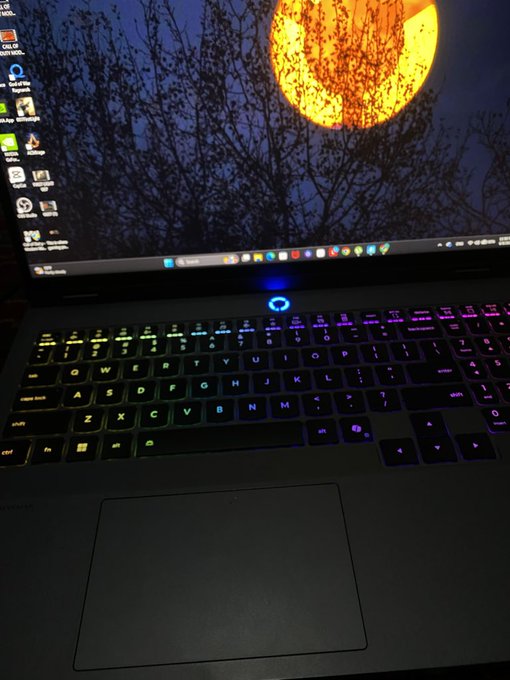 A laptop with a vibrant RGB-backlit keyboard displays a striking desktop wallpaper featuring a large orange moon behind silhouetted tree branches, with various application icons visible on the left side of the screen and a Windows taskbar at the bottom. The setup visually reinforces the post's message of "lightspeed" innovation and builder momentum, showcasing the developer's high-performance workstation after shipping their first AI-built appchain on the Canopy Network.