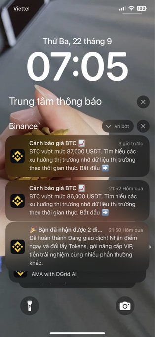 A smartphone lock screen showing Vietnamese text and time 07:05, with Binance notifications about BTC price alerts at $87,000 and $86,000, along with a daily check-in reminder.