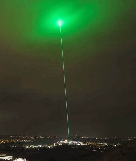 A night scene showing a bright green laser beam shooting into the sky from a cityscape, likely from a laser show or light installation.