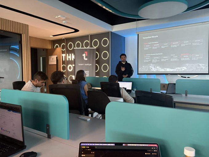 An instructor presents a 'Transaction lifecycle' slide to students seated at desks with laptops in a modern classroom featuring a 'team Turkiye' wall display.