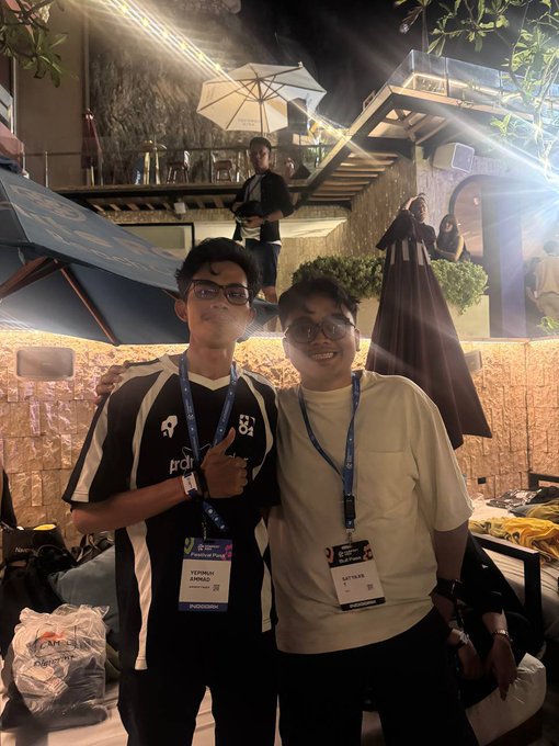 Two young men wearing lanyards and event passes, identified as 'YEYHAN ASHWAQ' and 'MITRA', posing for a photo at an indoor event space with an Ali B promotional poster in the background.
