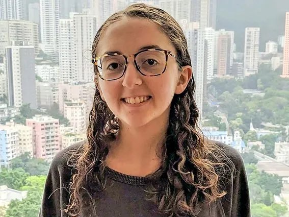 A young woman with curly brown hair and glasses smiles at the camera, with a hazy city skyline visible behind her. No text or identifying information is visible in the image itself, but the context suggests this may be Caroline Ellison, the former Alameda Research CEO who, according to the accompanying post, has been secretly working at the philanthropic platform Manifund under the pseudonym "Carol."