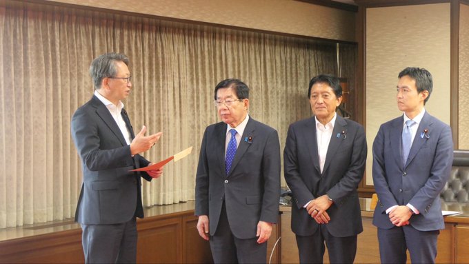 The image shows a group of four men dressed in suits, standing next to each other against a backdrop of a windowed room. One man appears to be speaking into a microphone or megaphone, while another man holds a red book. The fourth man is wearing a suit and tie, and the others are also dressed in formal attire. The scene suggests that they might be attending an event or gathering where they need to address an audience using the microphone or megaphone.