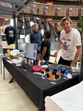 Three people stand behind a table displaying merchandise and accessories at what appears to be a pop-up event or vendor booth.