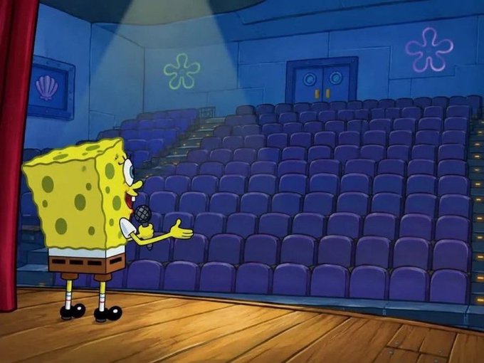 A SpongeBob SquarePants cartoon scene showing SpongeBob holding a microphone on stage, addressing an empty movie theater with blue seats and a spotlight overhead.