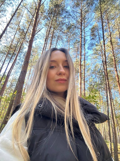 A selfie of a young woman with long blonde hair wearing a black puffer vest, standing in a sunny forest with tall trees.