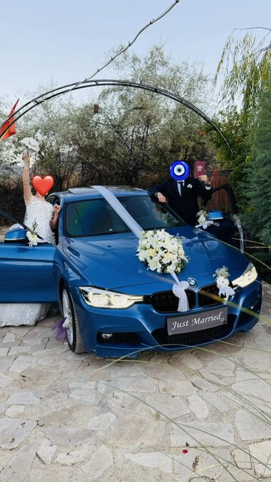 A blue BMW decorated with white ribbons and flowers for a wedding, featuring a 'Just Married' license plate, with a bride raising her arm and a groom waving beside the car.