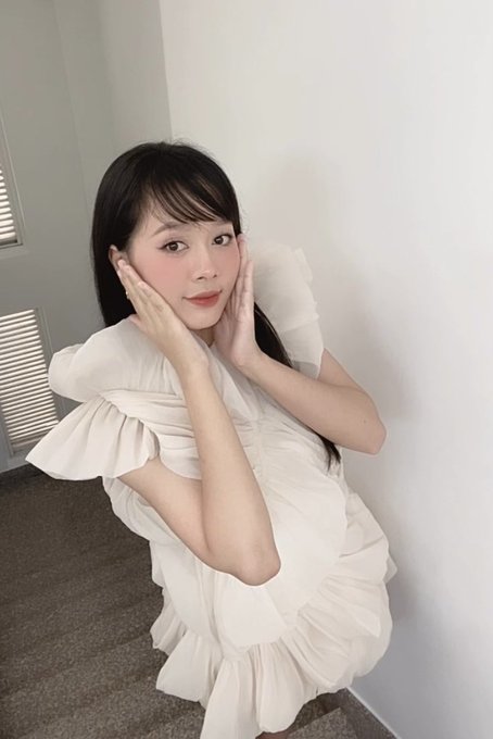 A young woman with long dark hair poses indoors, holding her cheeks with both hands while wearing a white, ruffled dress.