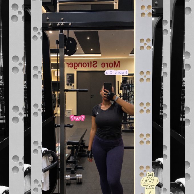 A gym mirror selfie showing a woman in a black athletic top and dark leggings holding a phone, with 'Mole Stronger' text (mirrored) visible on the glass and squat rack equipment surrounding her.