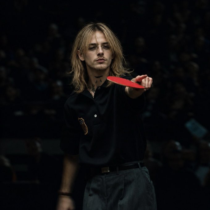 A man with long blonde hair and a goatee holds out a red ping pong paddle while wearing a black polo shirt in front of an audience.