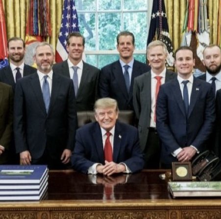 President Donald Trump is seated at the Resolute Desk with his hands clasped, surrounded by a group of men in suits standing behind him in the Oval Office, with American flags visible in the background and a row of blue binders placed on the desk.
