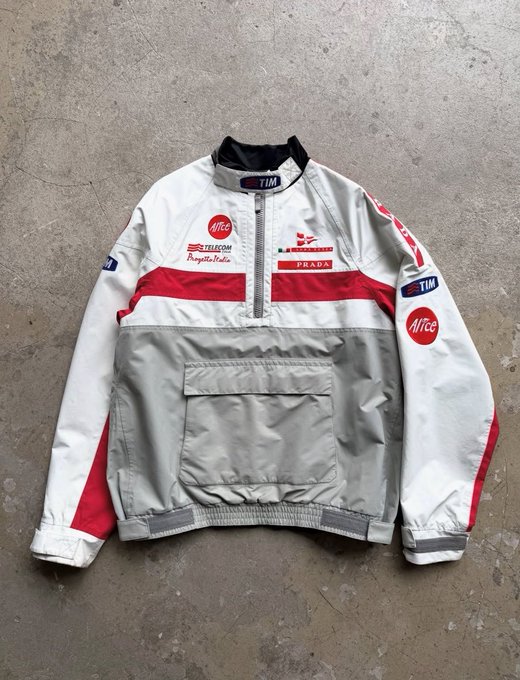 A white, gray, and red Prada Luna Rossa sailing team jacket laid flat on a concrete surface, featuring various sponsor logos including TIM, Pirelli, and Telefónica.