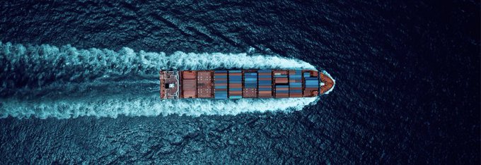 An aerial overhead photograph of a large container ship loaded with colorful shipping containers traveling through dark blue ocean waters.