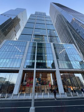A low-angle photograph captures a modern glass-and-steel skyscraper towering over a street-level entrance, with reflections of the surrounding city visible in the facade.