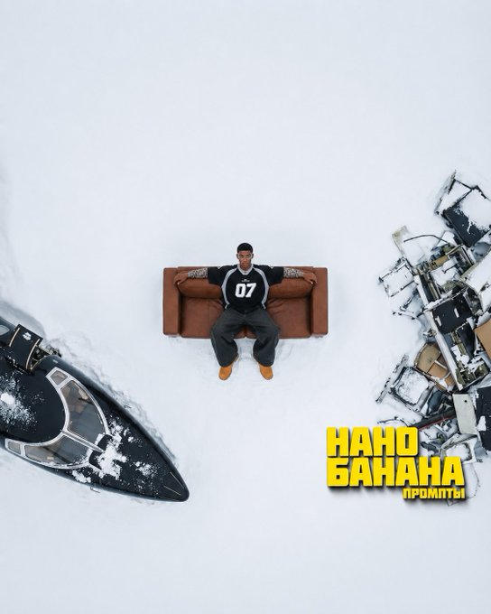A man in a black tracksuit and beanie with '07' on his chest, sitting on a brown couch in the snow, surrounded by a boat and debris, with the yellow text 'НАНО БАНА НА ПРОМИТЬ!' on the right.