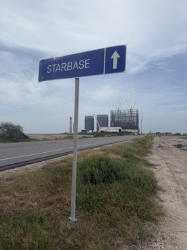 A blue road sign with a white arrow reads 'STARBASE' pointing toward SpaceX's Starbase facility visible in the distance along a rural road under an overcast sky.