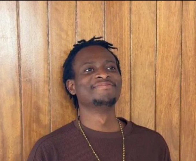 Casual portrait of a smiling young man with short dreadlocks and a light beard, wearing a maroon shirt and gold chain, standing in front of wood paneling.