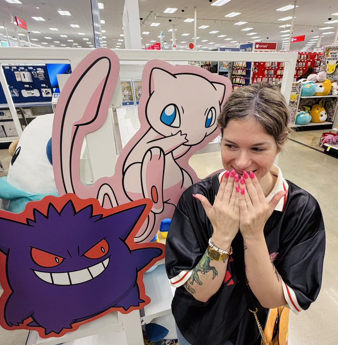 A woman with pink nails and tattoos poses excitedly with her hands near her face in a store, standing next to large cutouts of the Pokémon Mew and Gengar.