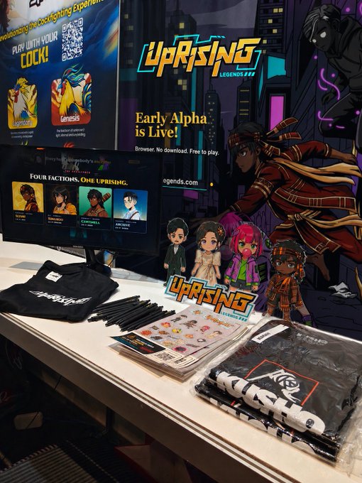 A promotional booth for "Uprising Legends" features a monitor showing "Four Factions. One Uprising," a banner stating "Early Alpha is Live!", and various merchandise including t-shirts, pens, stickers, and character standees. The display also includes branded apparel for "Kusho."