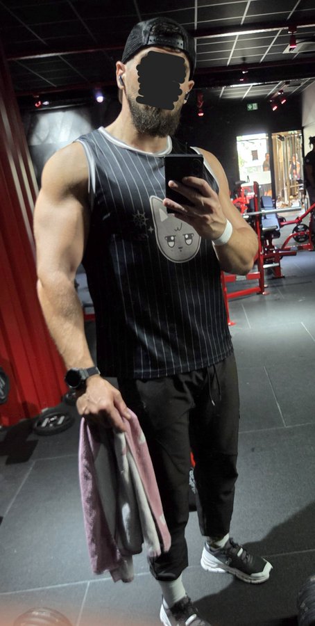 A muscular man wearing a black cap, a striped tank top with a cat face design, and black sweatpants takes a mirror selfie in a gym while holding a towel and cable attachment.