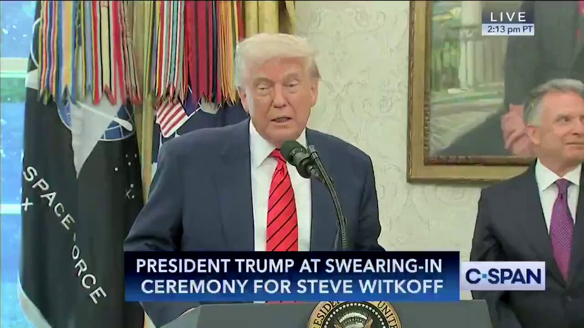 The image shows President Trump standing at a podium, wearing a suit with a red tie. He appears to be delivering a speech or addressing an issue. The background of the scene features multiple US flags hanging from the ceiling. There are also a couple of other individuals in the room who seem to be listening intently to the President's words. This suggests that it could be related to a significant event, announcement, or policy change as indicated by the caption provided.