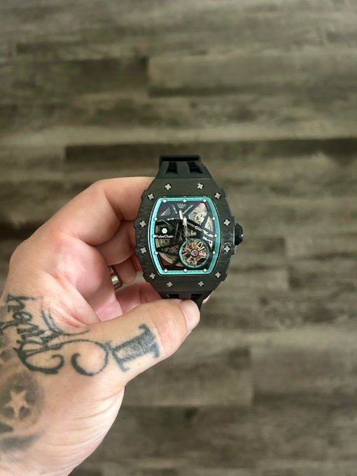 The image features a person's hand holding up a black Rolex watch with a blue face, showcasing its intricate design and craftsmanship. The background appears to be a wooden wall or floor, which complements the dark color of the watch. The caption "Focus on what is in the image and how it relates to the post #PulseChain" suggests that this image might be related to a social media post about watches or fashion accessories.