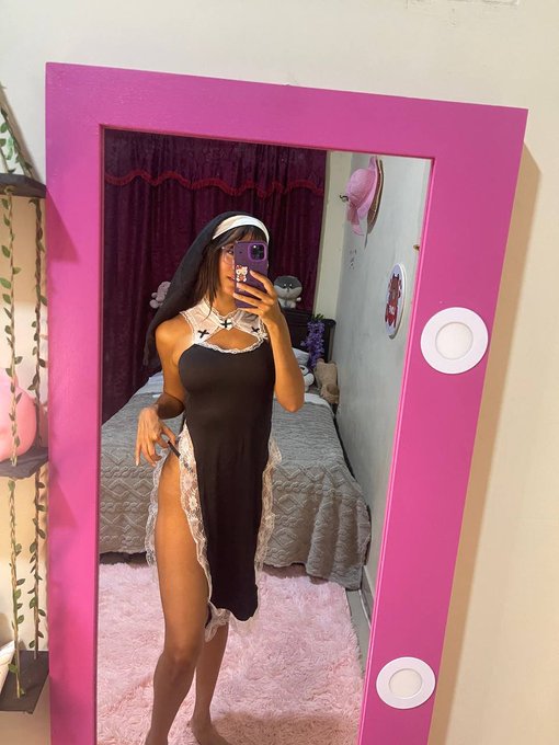 A woman takes a mirror selfie wearing a black dress and a white headpiece with a black veil, reflected in a mirror framed by a bright pink border.