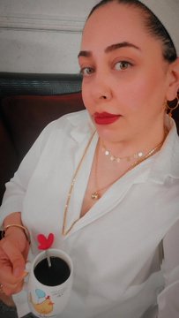 A woman wearing a white collared shirt and gold jewelry, holding a mug of dark coffee while seated on a brown leather couch.