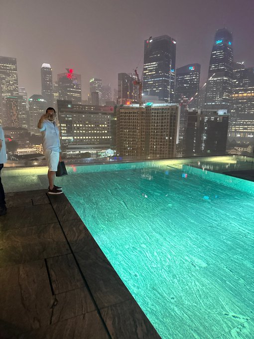 A person in white clothing stands by a glowing turquoise infinity pool overlooking a city skyline at night.
