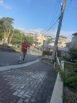 The image shows a man running down a brick sidewalk, wearing a red shirt and black shorts. The surrounding area includes buildings and power lines, indicating an urban setting. There are also two other people visible in the scene, one of whom appears to be carrying a backpack. The photo is taken from behind the runner, giving us a clear view of his actions as he runs down the street.

The post accompanying this image provides information about the man running and includes details such as the name of the person who took the picture (stepn) and the number of steps it takes to get there (stepngo). The photo also contains some text that reads "DIOさんのスペース聴きながらGARMINの 事故検知機能が働いてしまう".