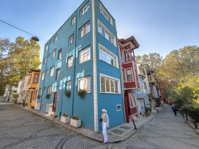 A narrow cobblestone alleyway lined with colorful, multi-story houses is shown with a woman walking in the foreground.