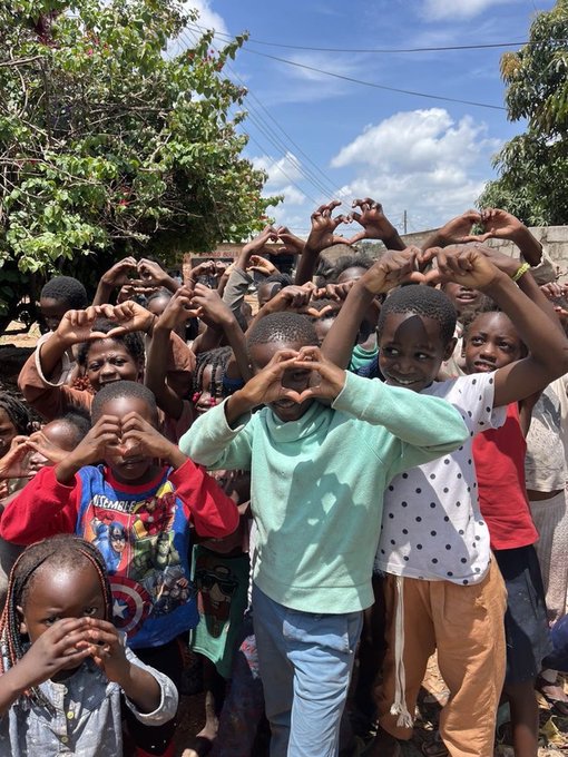 A group of children outdoors making heart shapes with their hands above their heads, smiling at the camera.