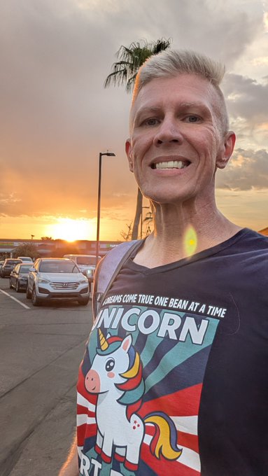 A smiling man with short grayish-blonde hair takes a selfie at sunset in a parking lot, wearing a dark t-shirt featuring a colorful unicorn graphic with the text 'UNICORN' and 'DREAMS COME TRUE ONE BEAM AT A TIME'. Palm trees and the ocean are visible in the golden background.