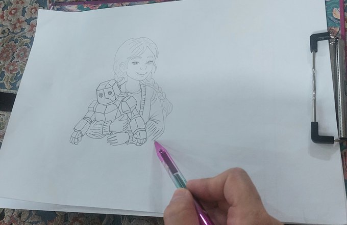 A hand holding a colorful pen is sketching a drawing of a young girl holding a robot on a white sketchpad clipped to a board.