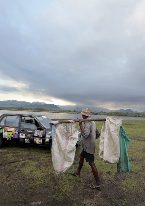 A man carries large white and teal sacks slung over a wooden pole across his shoulders, walking past a vehicle decorated with animal stickers (including pandas) on a marshy plain under an overcast sky. The image visually captures the "touching grass" theme of the post by depicting a rural, grass-grounded moment—though the identity of the passerby cannot be confirmed from the image alone, the playful tweet jokingly suggests it's Pumpfun founder @a1lon9 encountered unexpectedly in the wild.