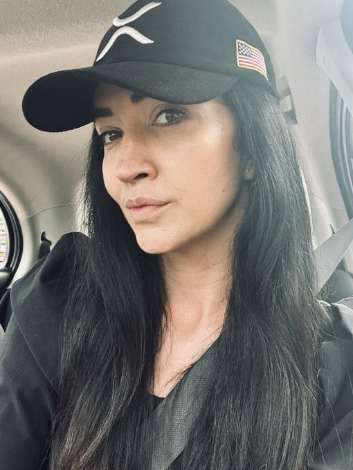 A selfie of a woman with long dark hair wearing a black X logo cap with an American flag patch, sitting inside a car.