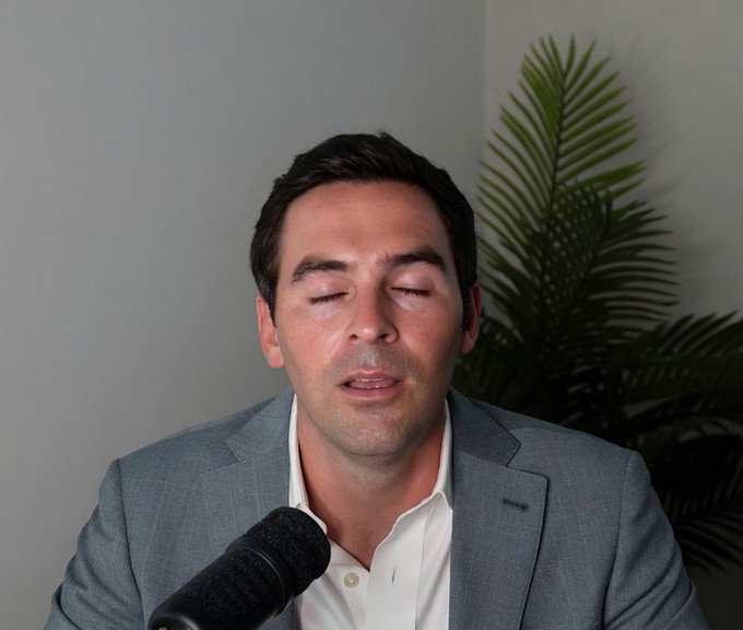 A man in a grey suit and white shirt speaking into a black microphone with his eyes closed, with a green plant in the background.