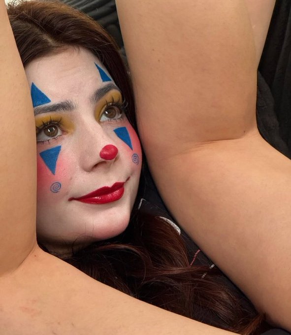 A woman with elaborate clown-style makeup featuring blue triangles, a red nose, and yellow eyeshadow poses with her arms raised above her head.