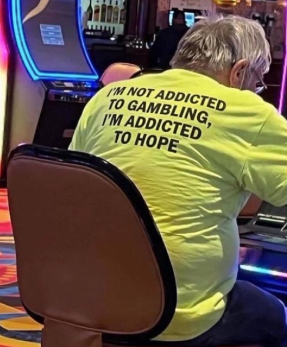 A person with blonde hair sits in a chair facing a slot machine, wearing a bright yellow t-shirt with the text 'I'M NOT ADDICTED TO GAMBLING, I'M ADDICTED, TO HOPE' printed on the back.