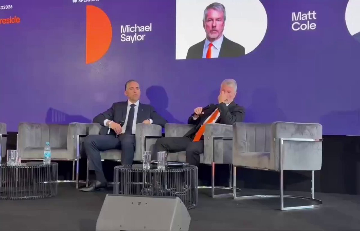 The image shows two men sitting in chairs at a conference room with a purple wall behind them. One man is wearing a suit and tie while the other has his legs crossed, appearing relaxed. The background features a large screen displaying an interview with Michael Saylor talking about Ethereum's collapse due to competition from Solana, BNB Chain, and other L2s.