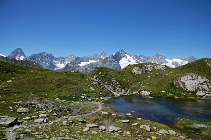 A scenic mountain landscape with snow-capped peaks under a clear blue sky, a small alpine lake, and grassy terrain in the foreground.