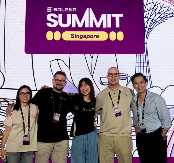 The co-hosts of Solana Summit Singapore pose together beneath the event's signage, which displays the "SOLANA SUMMIT Singapore" branding in purple and yellow. The group of five attendees, all wearing event lanyards, smiles for the camera in front of the illustrated backdrop, capturing the collaborative spirit of the gathering that drew 2,048 attendees from 93 countries.