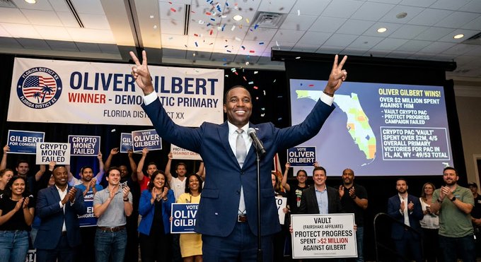 Oliver Gilbert stands triumphantly with both arms raised in victory at a campaign event, celebrating his win over a $2 million crypto industry primary campaign, as displayed on the screen reading "Oliver Gilbert Wins Over $2 Million Spent Against Him" and "Protect Progress Spent ~$2 Million, Crypto PAC Spent Over $2M, Overall Primary Victories for PAC in FL, AK, WV - 49/53 Total." Supporters behind him hold signs reading "Crypto Money Failed" and "Oliver Gilbert," illustrating the unsuccessful Fairshake and Protect Progress efforts described in the accompanying post.