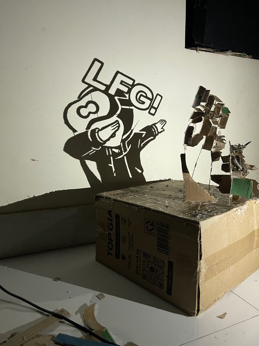 The image features a cardboard box with a drawing of a person on it, which says "LEG!" above their head. The box appears to be sitting on top of a white surface or wall. There are also some tape and other items scattered around the scene, including a computer mouse near the bottom right corner and a green item in the middle left area.