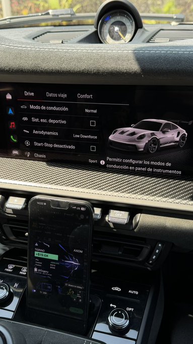 The dashboard of a Porsche displays driving settings in Spanish ("Drive," "Datos viaje," "Confort") alongside an image of a sports car, while a smartphone rests on the center console charging via a $19.00 Energy service. The image perfectly embodies the "low cortisol weekend" vibe—a quiet, mindful pause in a luxury vehicle's cabin, blending technology and tranquility.