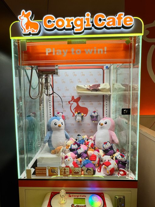 A "Corgi Cafe" claw machine labeled "Play to win!" is filled with Pudgy Penguins plushies alongside larger penguin characters, promoting the brand at a high-traffic intersection of tech, AI, finance, and culture that's open 24/7.