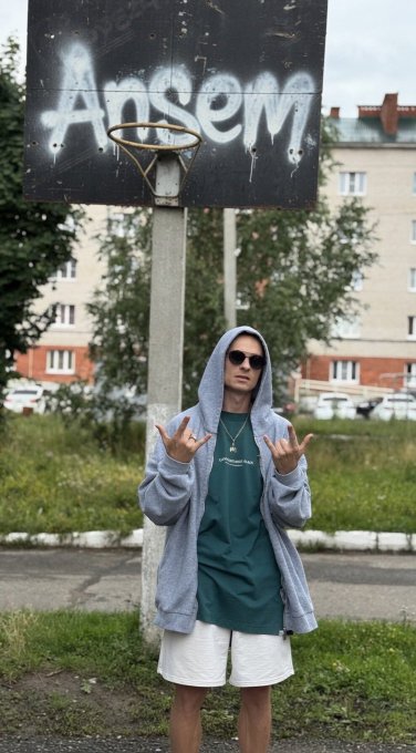 A young man in a gray hoodie, green shirt, and sunglasses poses under a weathered basketball backboard spray-painted with "ANSEM," flashing hand gestures in a Russian residential neighborhood—visually reinforcing the post's joke that the $ANSEM meme community has spread globally, even reaching Russia.