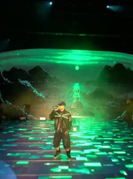 A child performs on a stage bathed in green lighting, with a digital LED floor and a backdrop depicting a moonlit landscape.